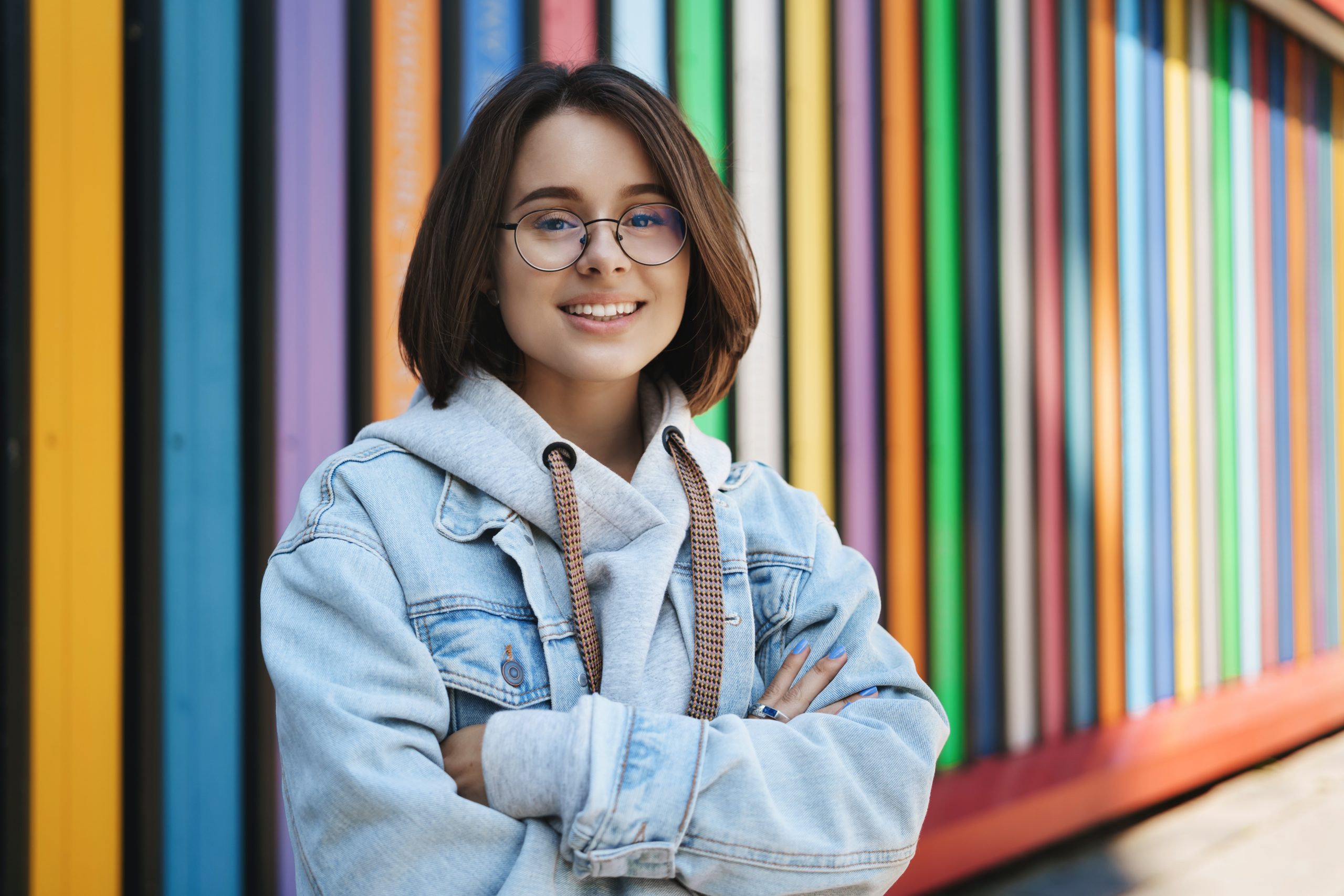 Cheerful young queer girl in glasses, denim jacket, standing near rainbow wall, cross hands over chest and smiling camera happy, lifestyle, urban life and generation concept.